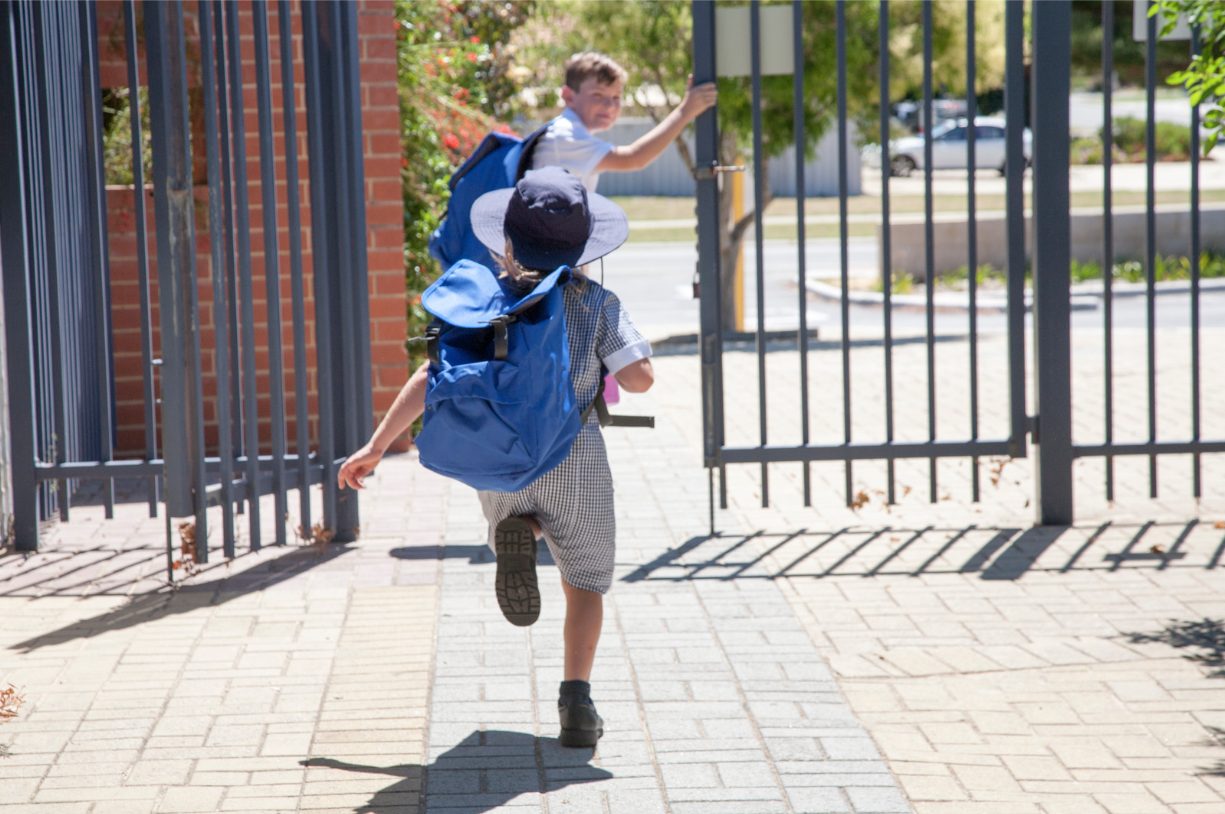 A child in a sunhat and blue backpack running through the school gate, another child holding it open