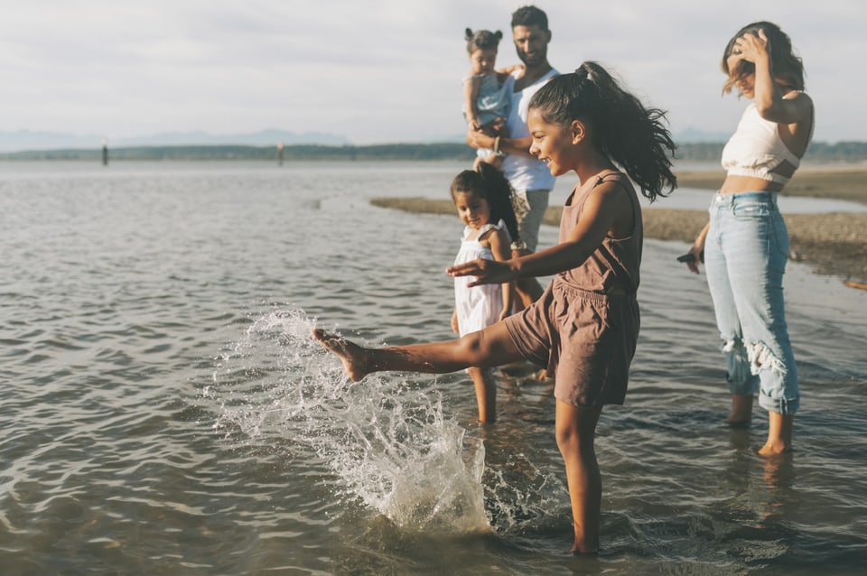 A family paddling at the edge of the water, a girl kicking up a splash while her parents and sisters watch