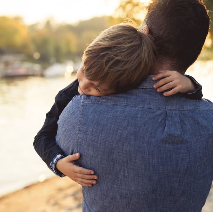 A young boy resting his head on his father's shoulder in a hug, by the water at sunset