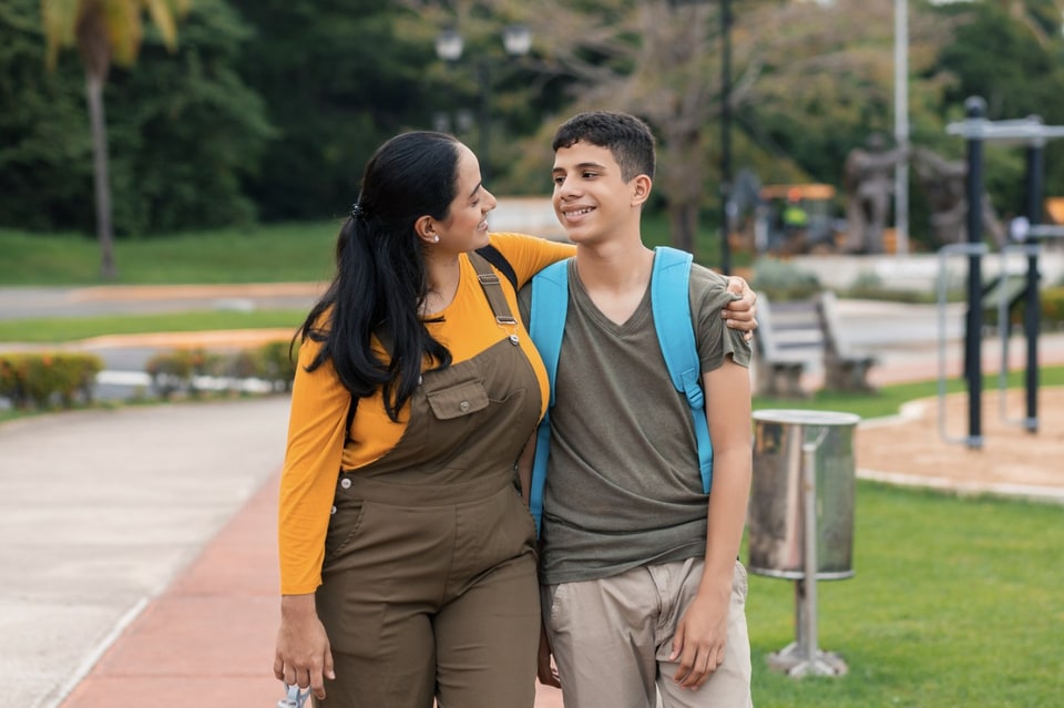 A parent walking arm in arm with their teenage son on the way to school, both smiling